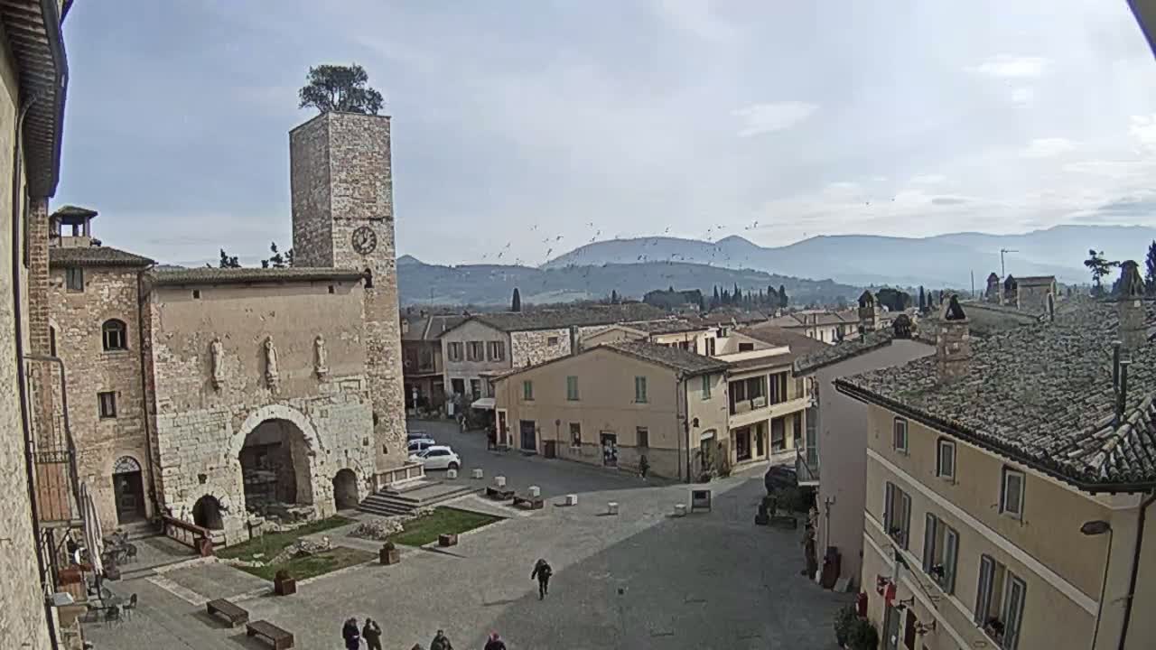 A town square with stone buildings, a clock tower with a tree on top, and a view of distant mountains under a mostly clear sky.