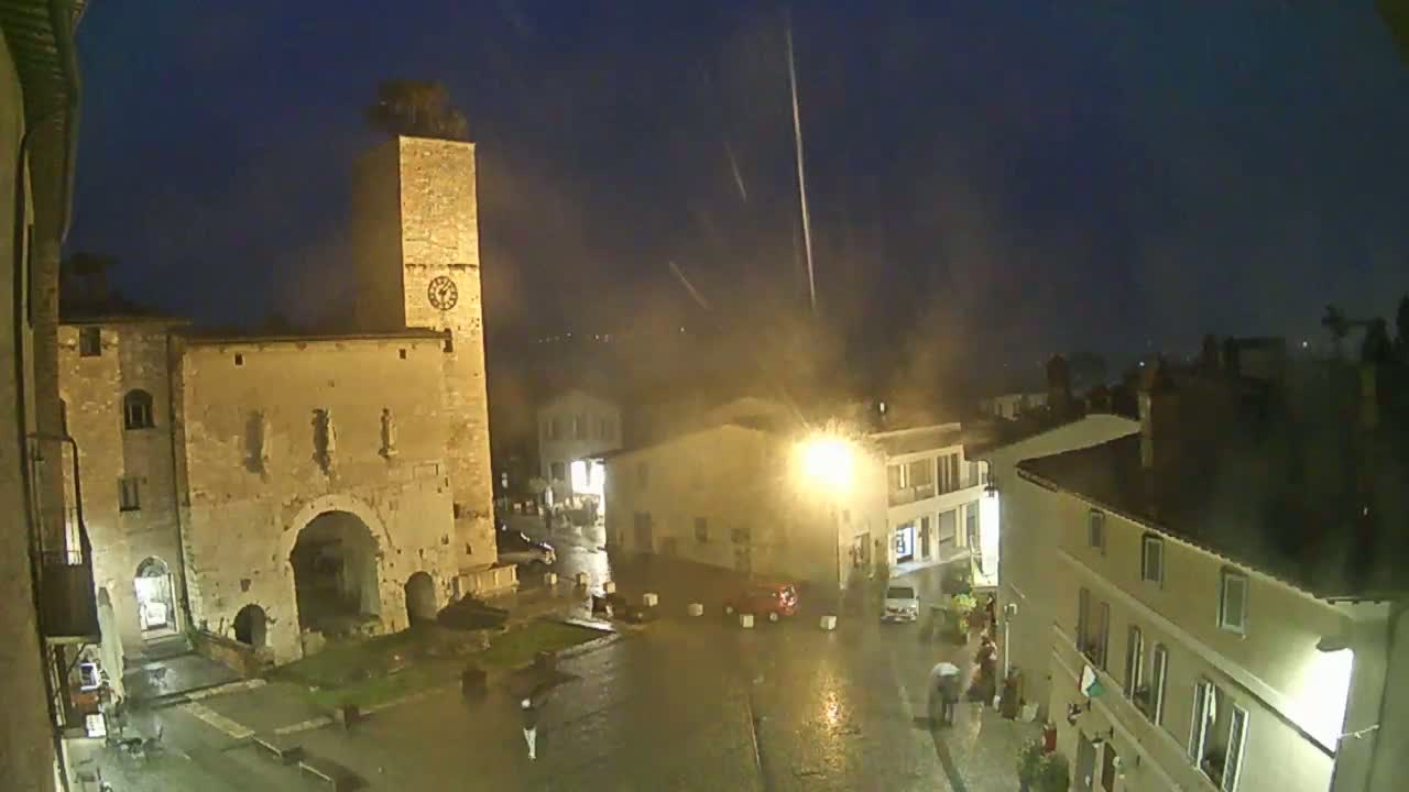 A nighttime view of a town square with a large stone building and clock tower, under a rain shower.