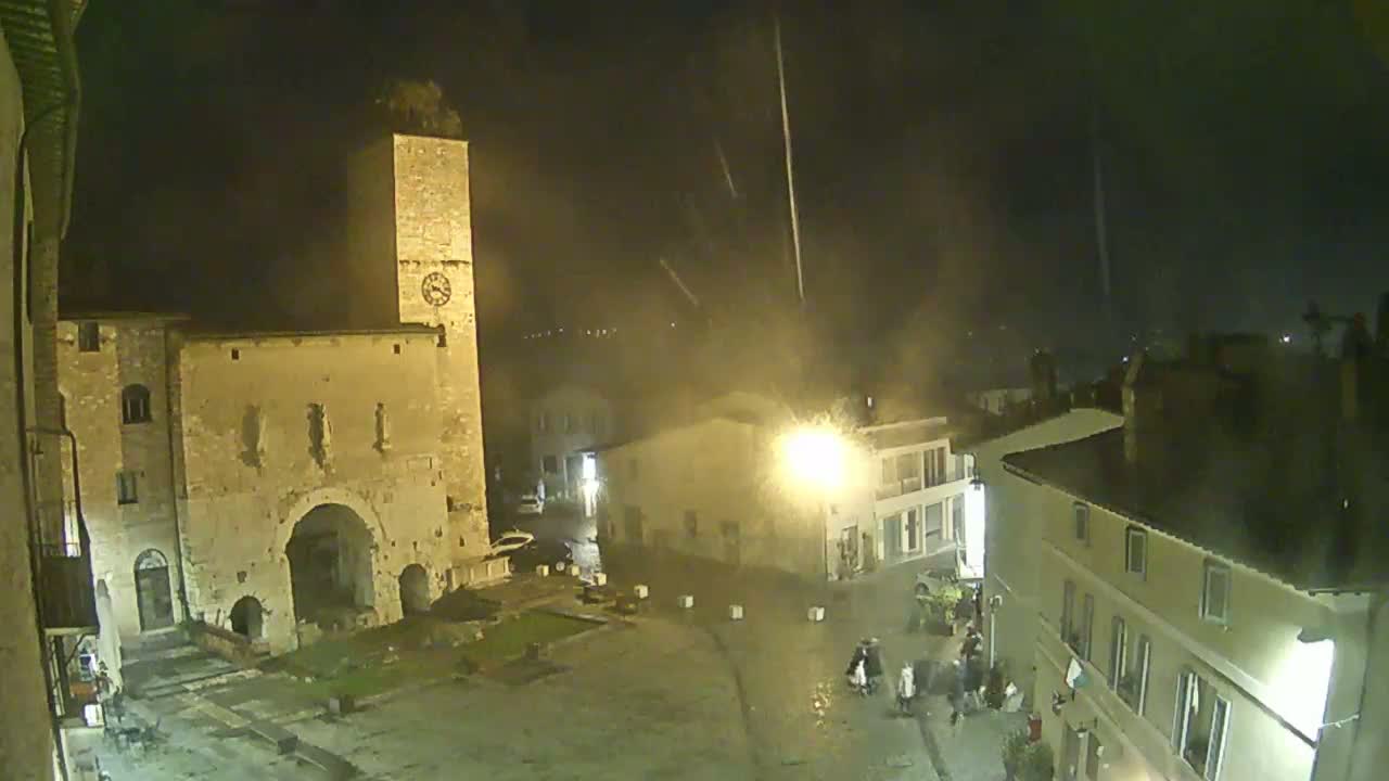 A nighttime, rain-swept view of a town square, featuring an old stone building with a clock tower and several other buildings under dim lighting.