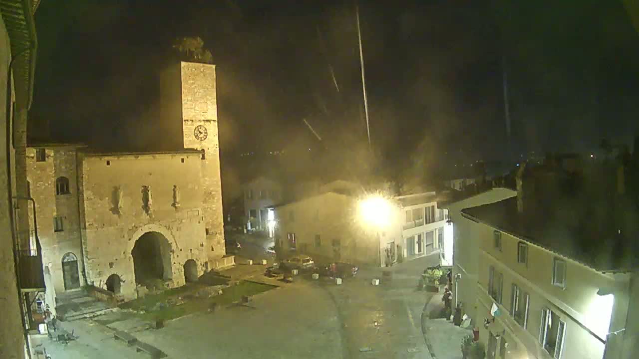 A nighttime view of a town square with old stone buildings, a tall clock tower, and light rain falling.