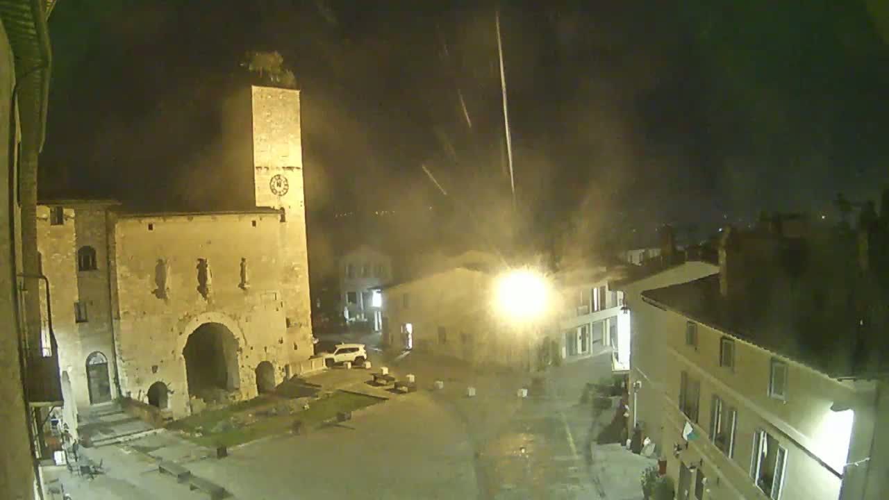 A nighttime view of a town square, featuring an old stone building with a clock tower, other buildings, and a car, under a light rain or drizzle.