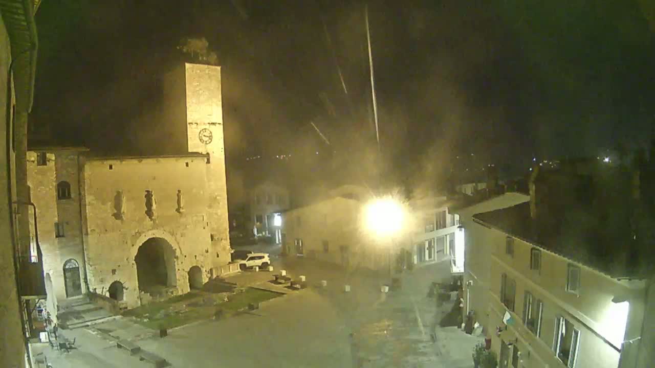 A nighttime, possibly rainy, view of a town square with ancient stone buildings, a clock tower, and a few cars parked nearby.