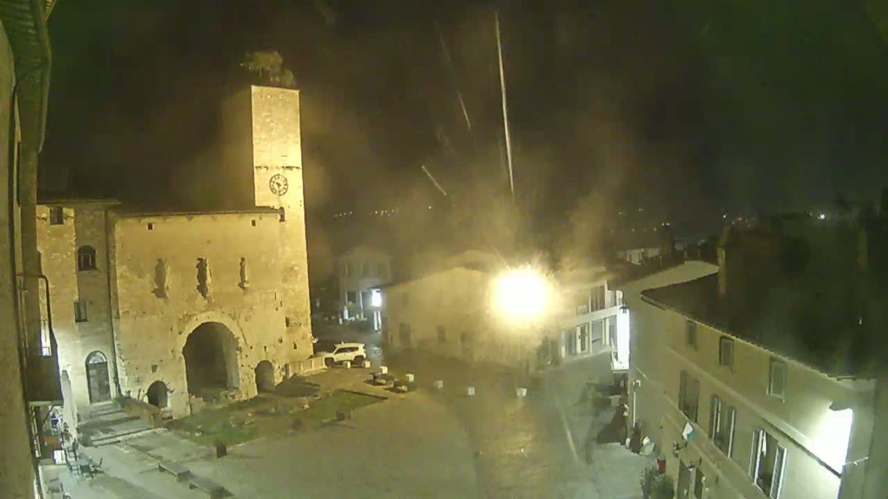 A nighttime view of a town square, featuring an old stone tower and buildings under hazy, possibly rainy conditions.