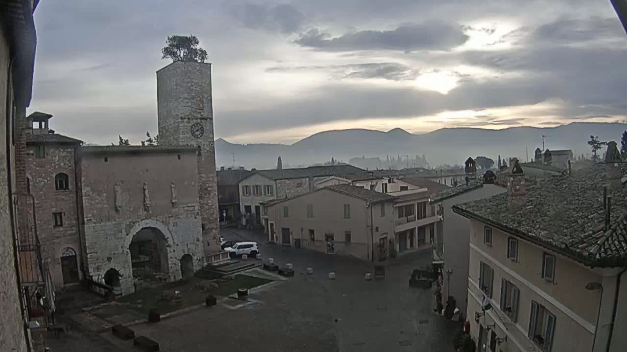 A town square with stone buildings, a clock tower topped with a tree, and distant hazy mountains under a cloudy, sunrise sky.