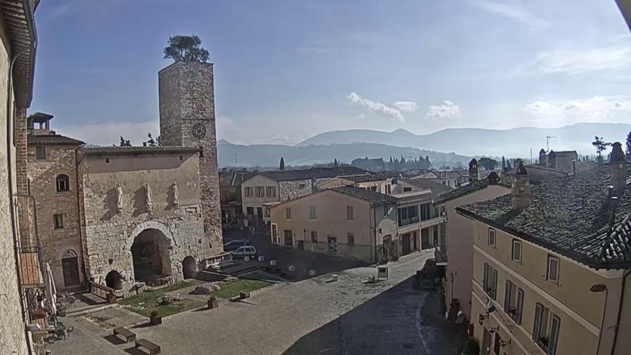 A town square with a clock tower and stone buildings, under a mostly clear sky with distant mountains visible in the background.