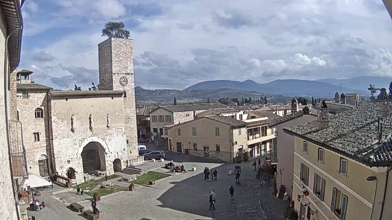 A town square with a stone tower, buildings, and people, under a partly cloudy sky with mountains in the background.