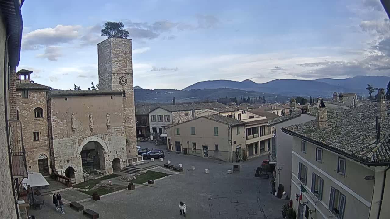 A town square with stone buildings, a clock tower topped with a tree, and distant mountains under a partly cloudy sky.