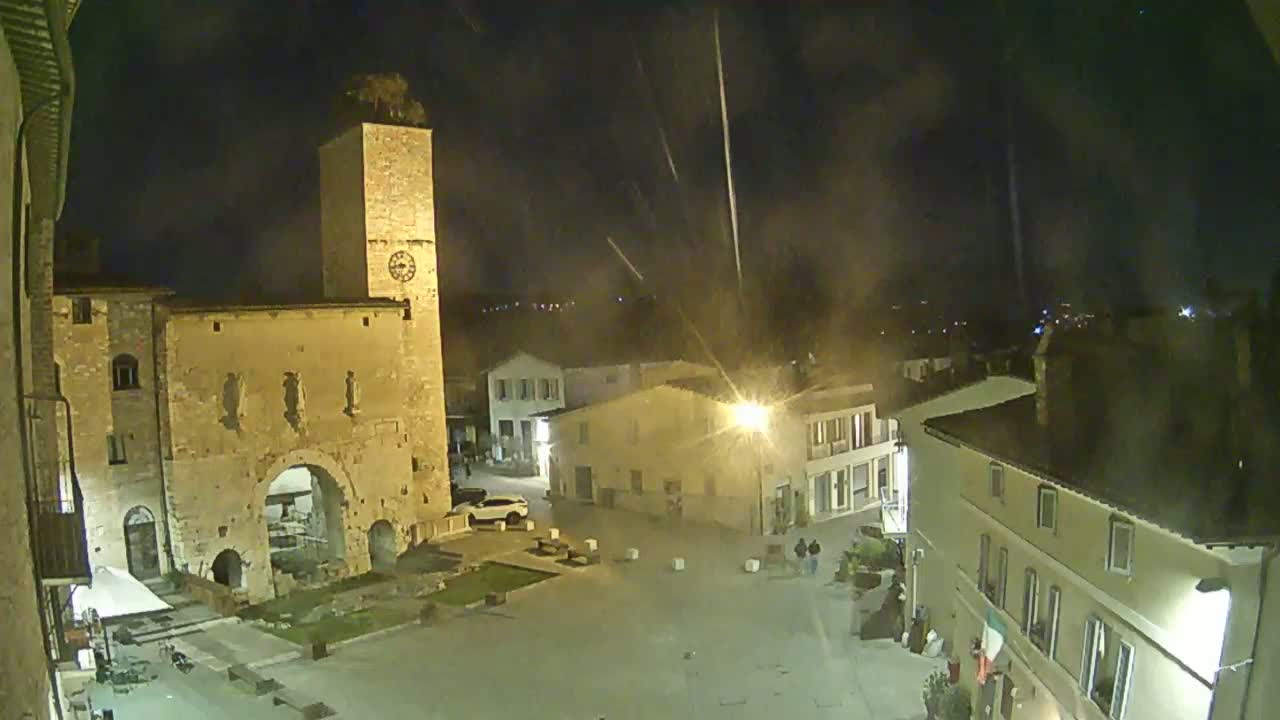 A nighttime view of a town square with an ancient stone gate and clock tower, under a slightly hazy sky with streaks of light possibly from fireworks or aircraft.