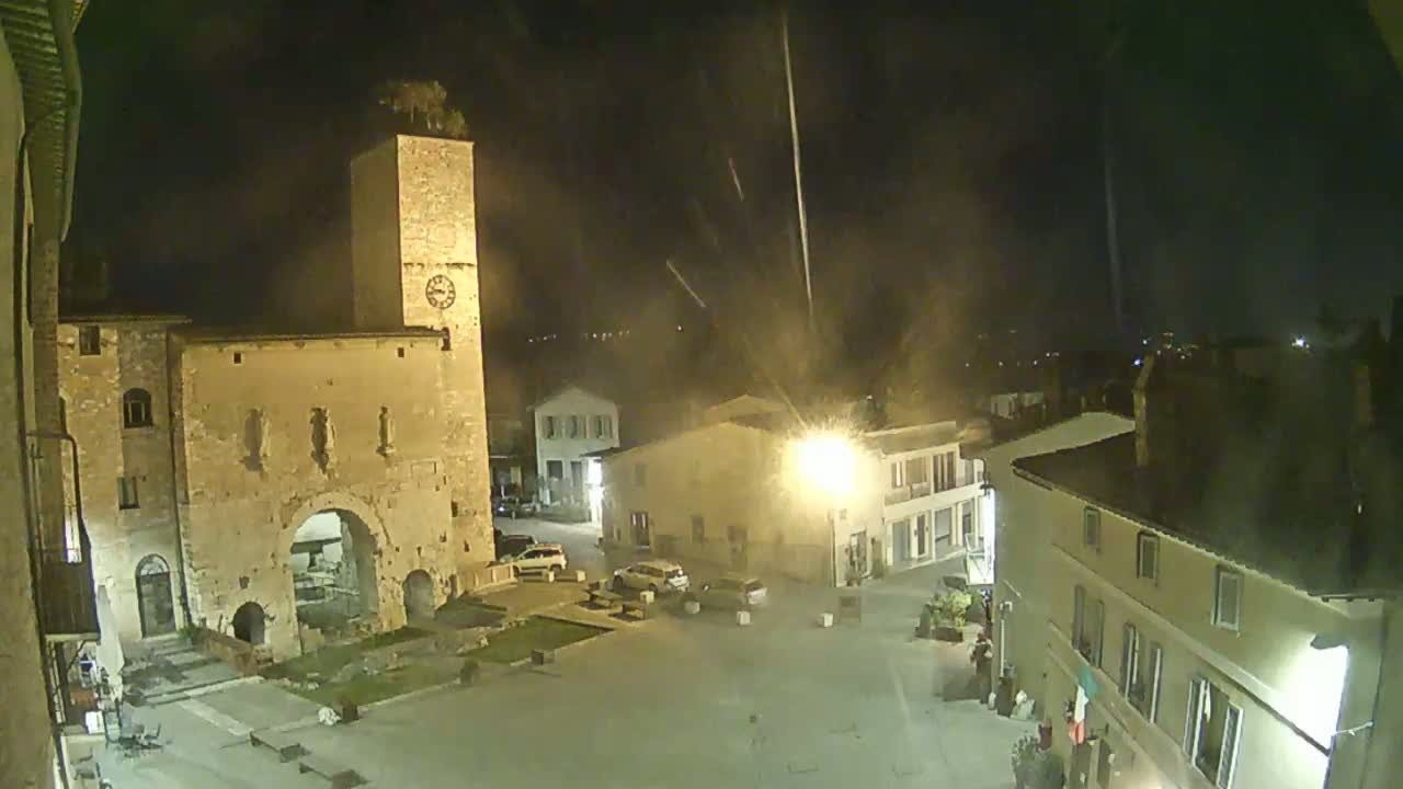 A nighttime view of a town square with stone buildings, including a tall clock tower, and a few cars parked nearby under a hazy sky.