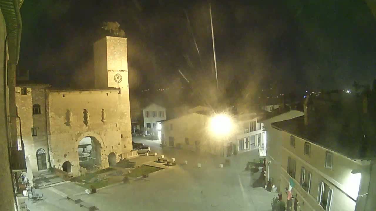 A nighttime view of a town square, featuring an old stone gate and clock tower, under a hazy sky with streaks of light possibly from rain or fireworks.