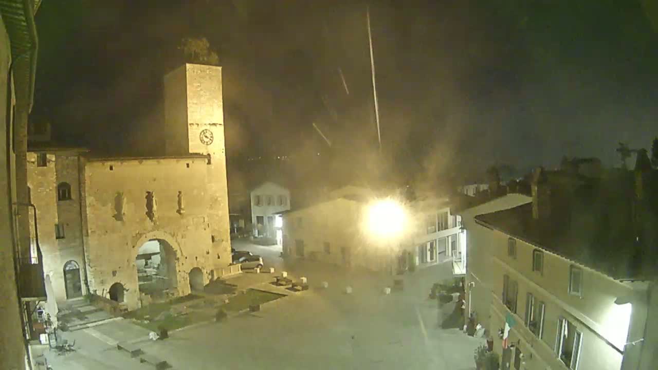 A nighttime, high-angle view shows an open town square with a tall stone tower and several buildings illuminated under hazy, possibly misty, conditions.
