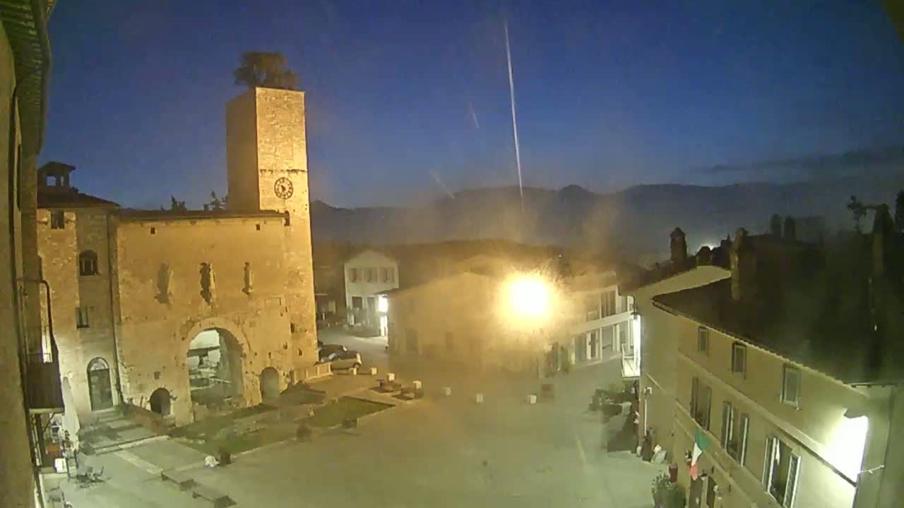 A nighttime, slightly hazy view of a town square dominated by a tall stone tower with a clock, surrounded by illuminated buildings.