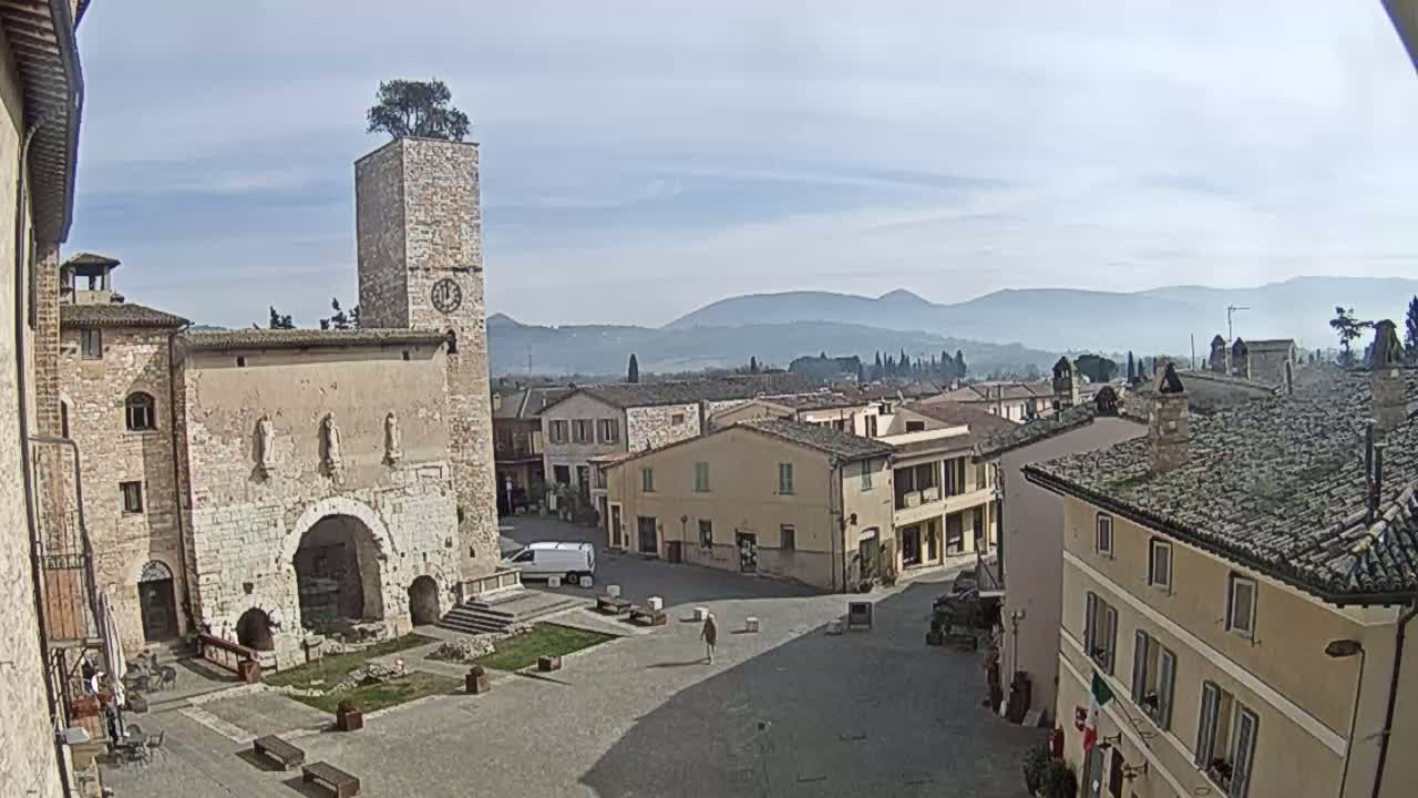 A town square in a small Italian town, under a partly cloudy sky, is surrounded by old stone buildings, one with a clock tower topped by a tree.