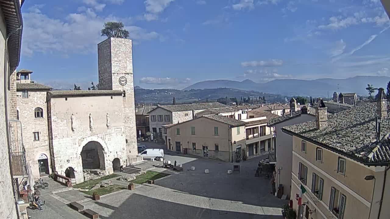 A mostly sunny day reveals a town square in a small Italian town, featuring a clock tower with a tree on top, surrounded by stone buildings and overlooking distant mountains.