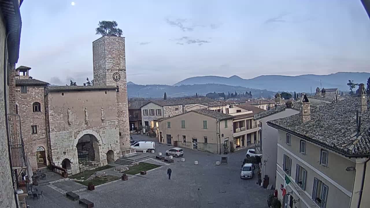 A town square in a small, historic town, under a pale, partly cloudy sky, is surrounded by stone buildings and overlooks distant mountains.