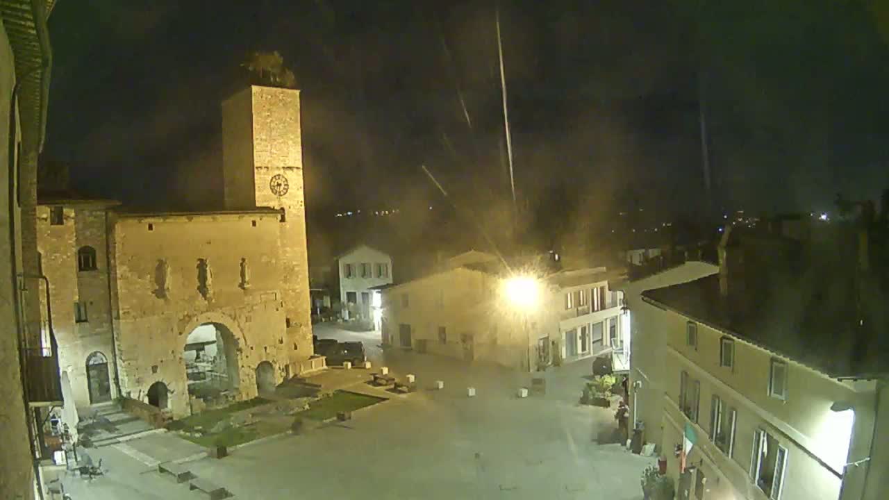 A nighttime view of a town square, illuminated by streetlights, features an ancient stone structure with a tall tower and surrounding buildings under a hazy sky with streaks of light.