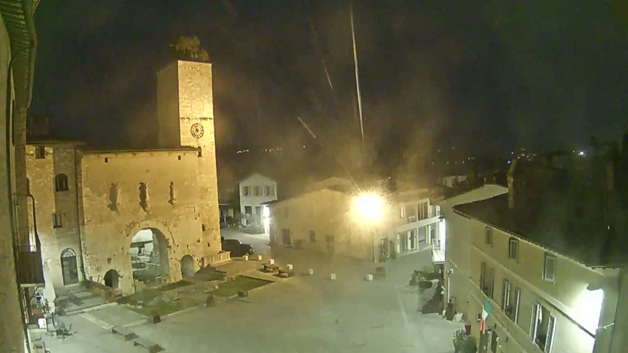 A nighttime view of a town square with a stone tower and buildings, under hazy conditions with what appears to be fireworks in the sky.