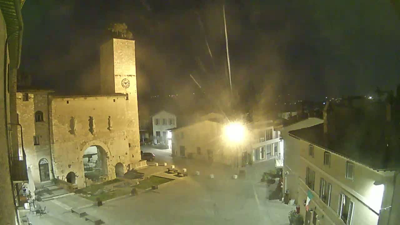 A nighttime view of a town square with an old stone gatehouse and clock tower under a slightly hazy sky.