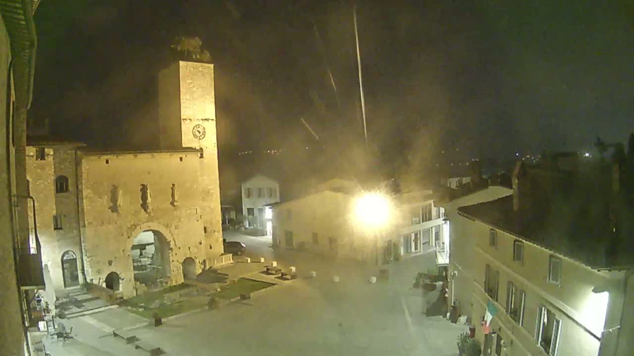 A nighttime view of a town square with a tall stone tower and several buildings under a hazy, possibly rainy sky.