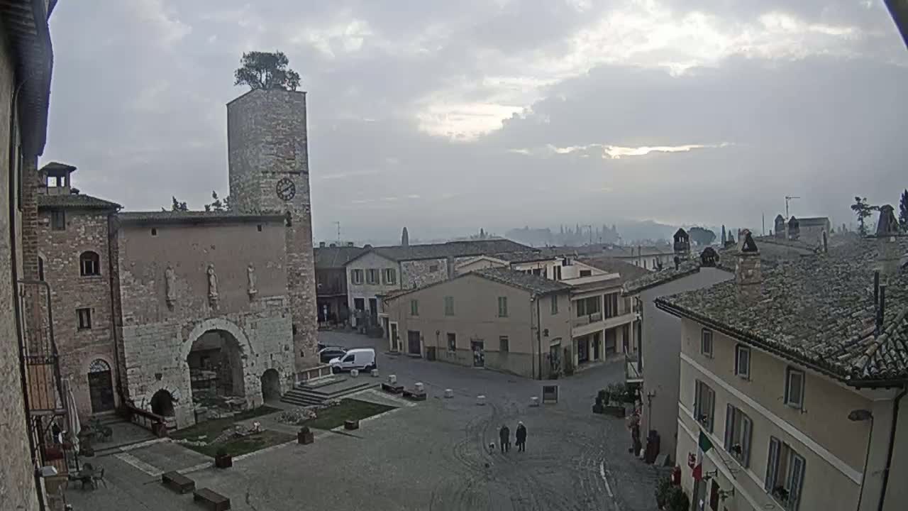 A town square with stone buildings, a clock tower topped with a tree, and a few people walking under an overcast sky.