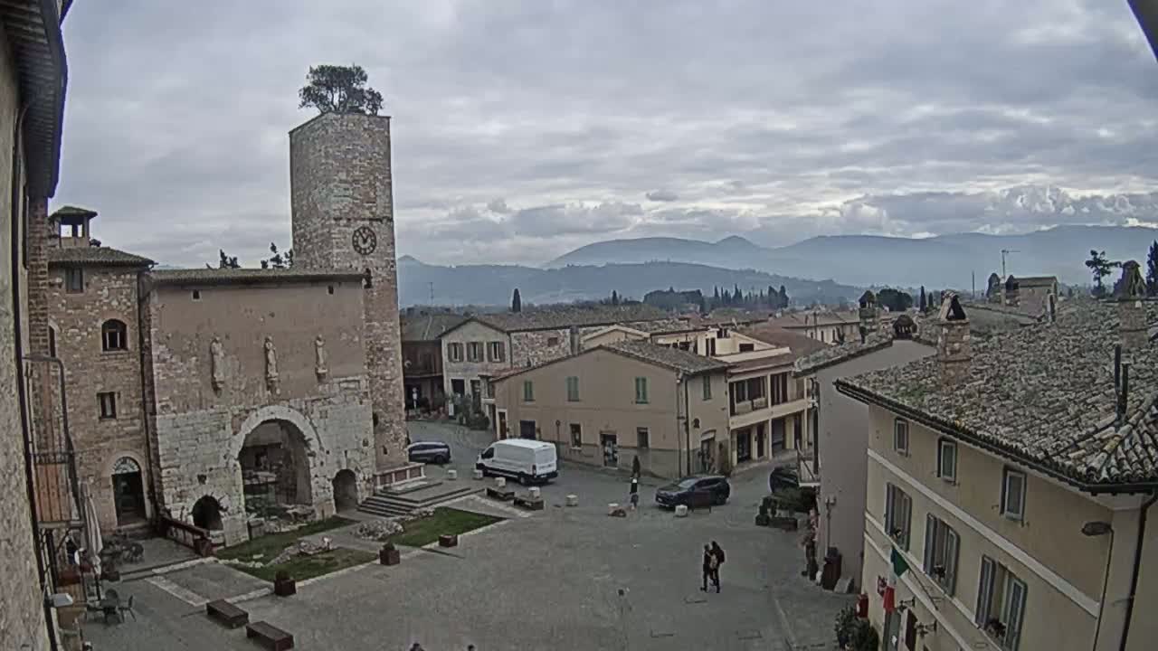 A town square in a historic town, under a cloudy sky, is surrounded by stone buildings, one with a tall clock tower topped with a tree, and mountains are visible in the distance.