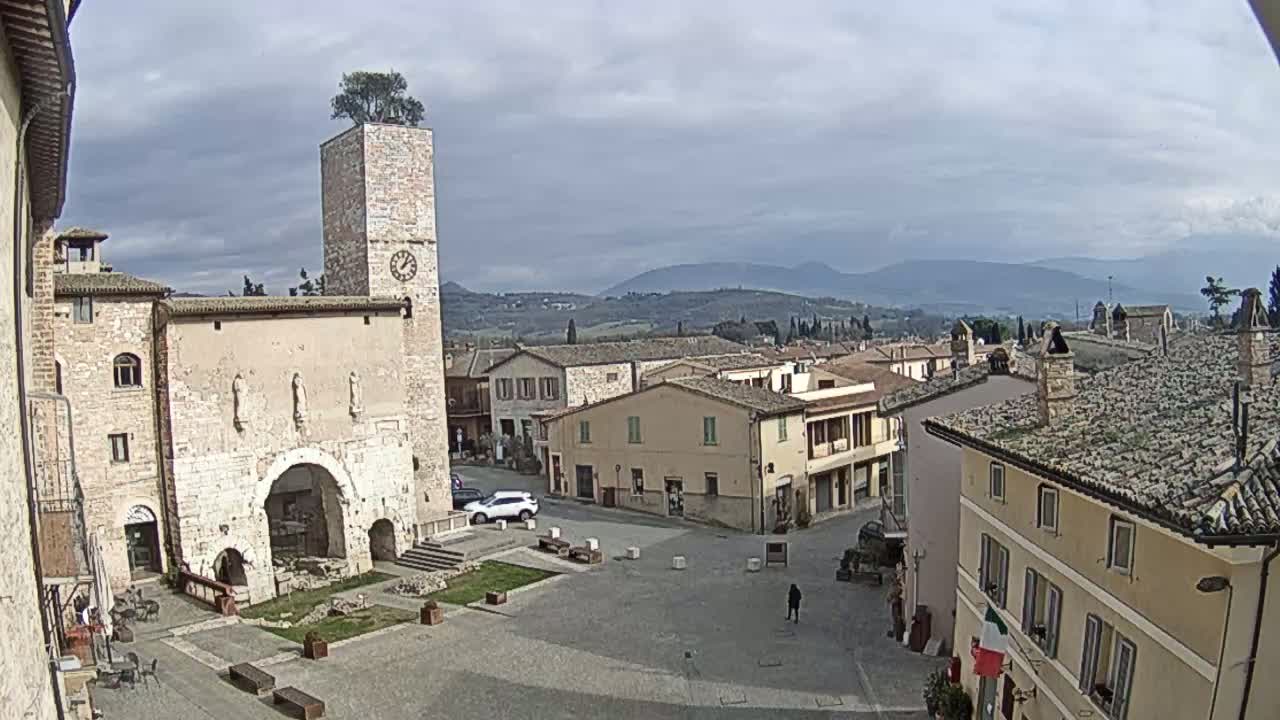 A town square in a small Italian town, under a cloudy sky, is surrounded by stone buildings, one of which has a clock tower with a tree on top.