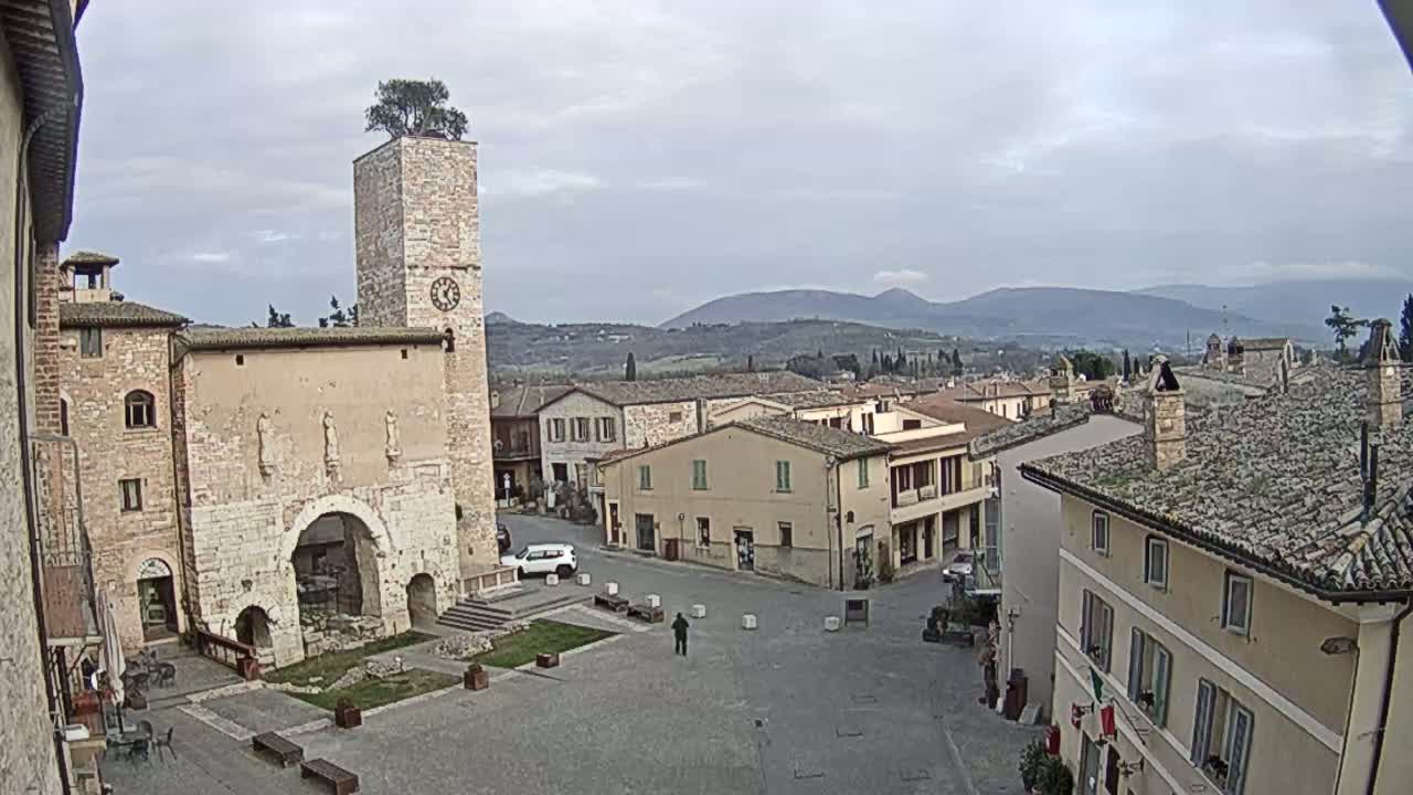 A town square in a small Italian town, under an overcast sky, is surrounded by stone buildings, one of which has a tall clock tower with a tree on top, and overlooks rolling hills in the distance.