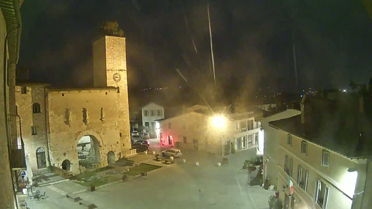 A town square at night, illuminated by streetlights, shows a stone tower and buildings under light rain.