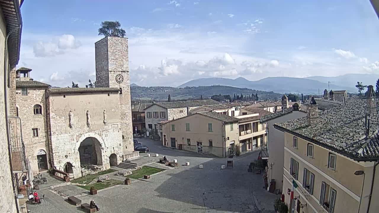 A town square in a village nestled in a valley, under a mostly sunny sky with some clouds, is surrounded by buildings of varying heights, with a prominent clock tower featuring a tree on its top.