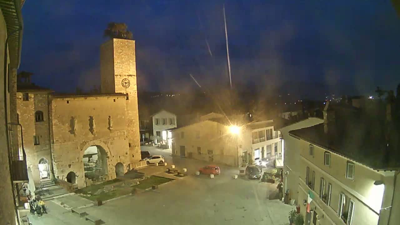 A nighttime view of a town square, featuring a tall stone tower with a clock, surrounding buildings illuminated by streetlights, and a few cars parked, under slightly hazy conditions.