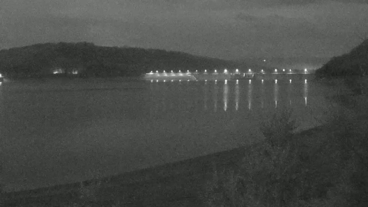 At night, a body of water reflects the lights of a structure across its surface, with dark hills visible in the background under a dark sky.