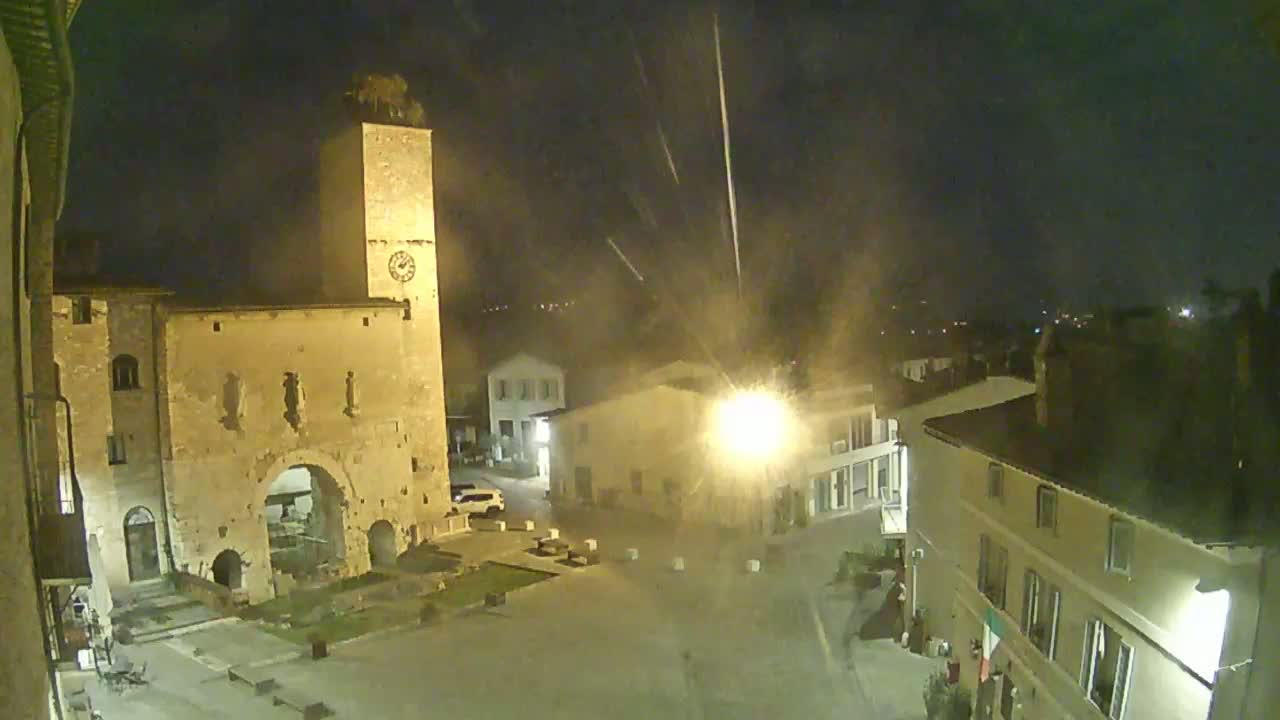 A nighttime view of a town square, dominated by an old stone building with a clock tower, under conditions of light rain or drizzle.