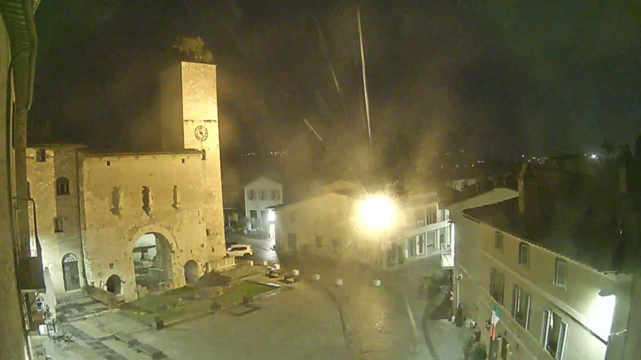 A nighttime view of a town square with an old stone building and tower under a light rain.