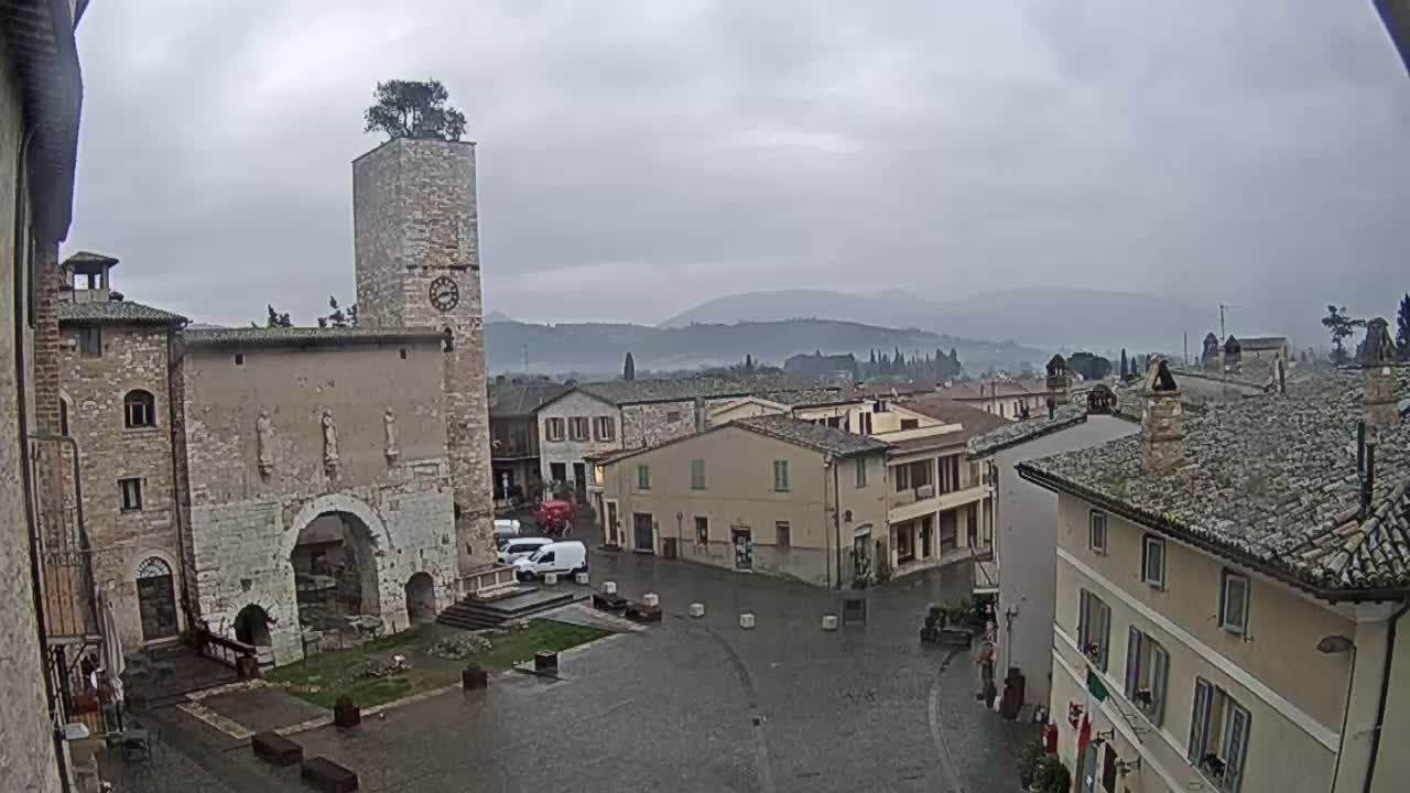 A rain-soaked town square in a hilly region, centered on a stone building with a clock tower and archway, features several buildings of similar architecture, under an overcast sky.