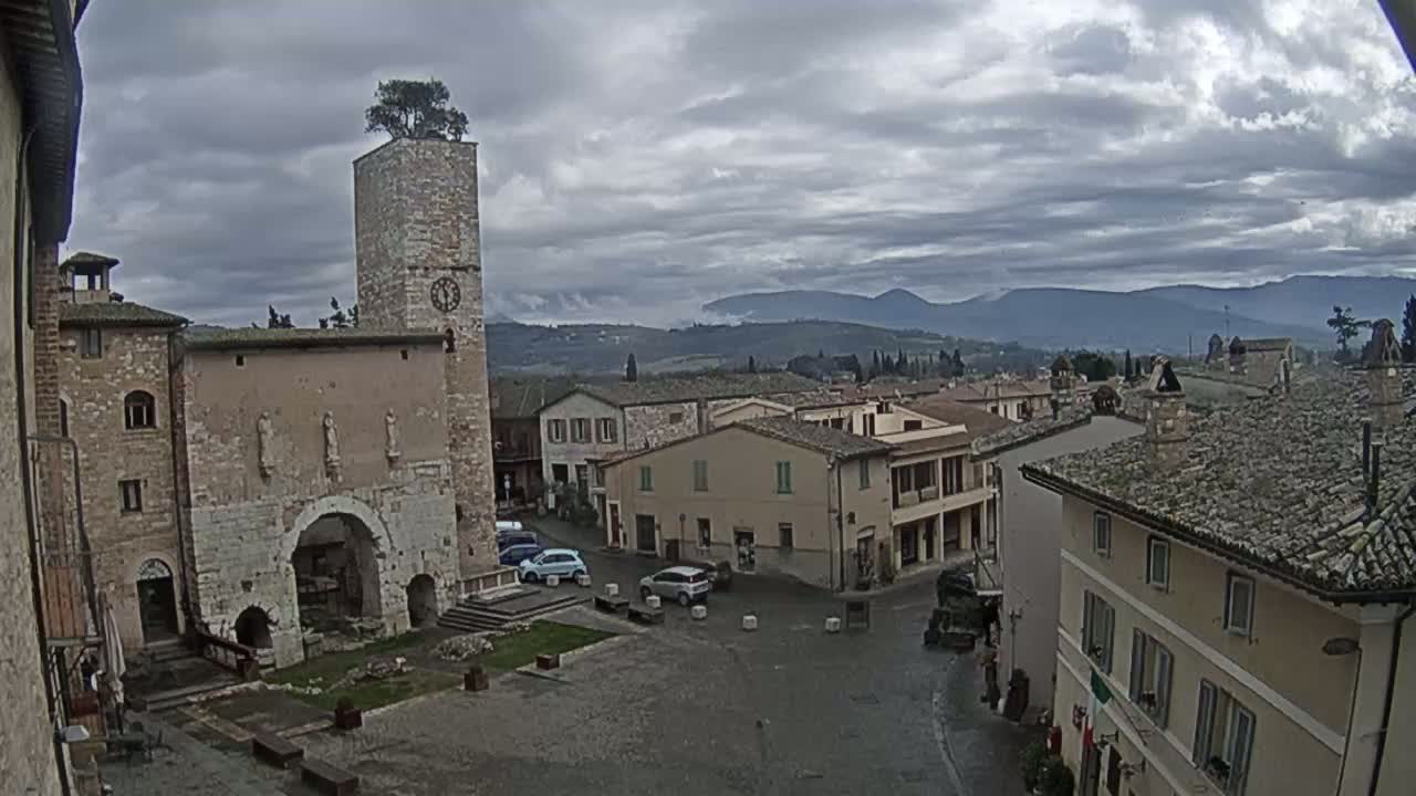 A town square with several stone buildings, a clock tower with a tree on top, and a few cars, under a cloudy sky with distant mountains visible in the background.