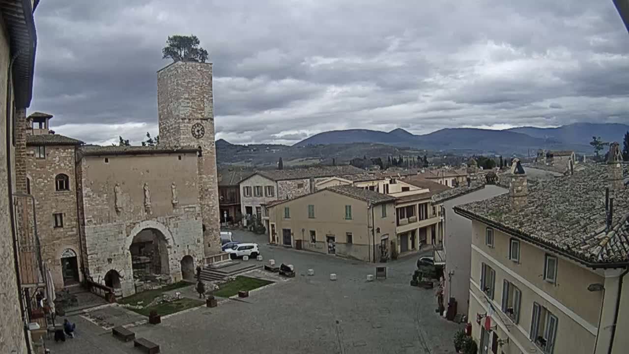 A town square with old stone buildings, a clock tower topped with a tree, and distant mountains under a cloudy sky.