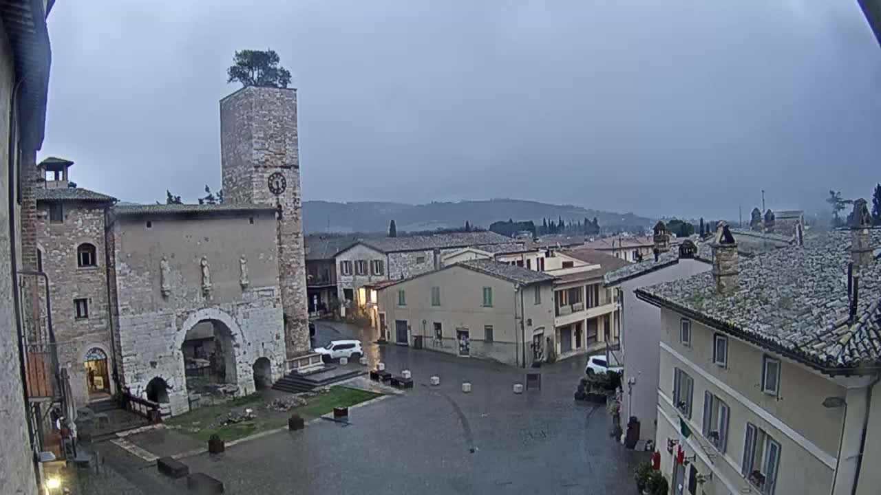 A rain-slicked town square in a medieval-style town, featuring a tall stone tower with a tree on top, is surrounded by buildings under an overcast sky.
