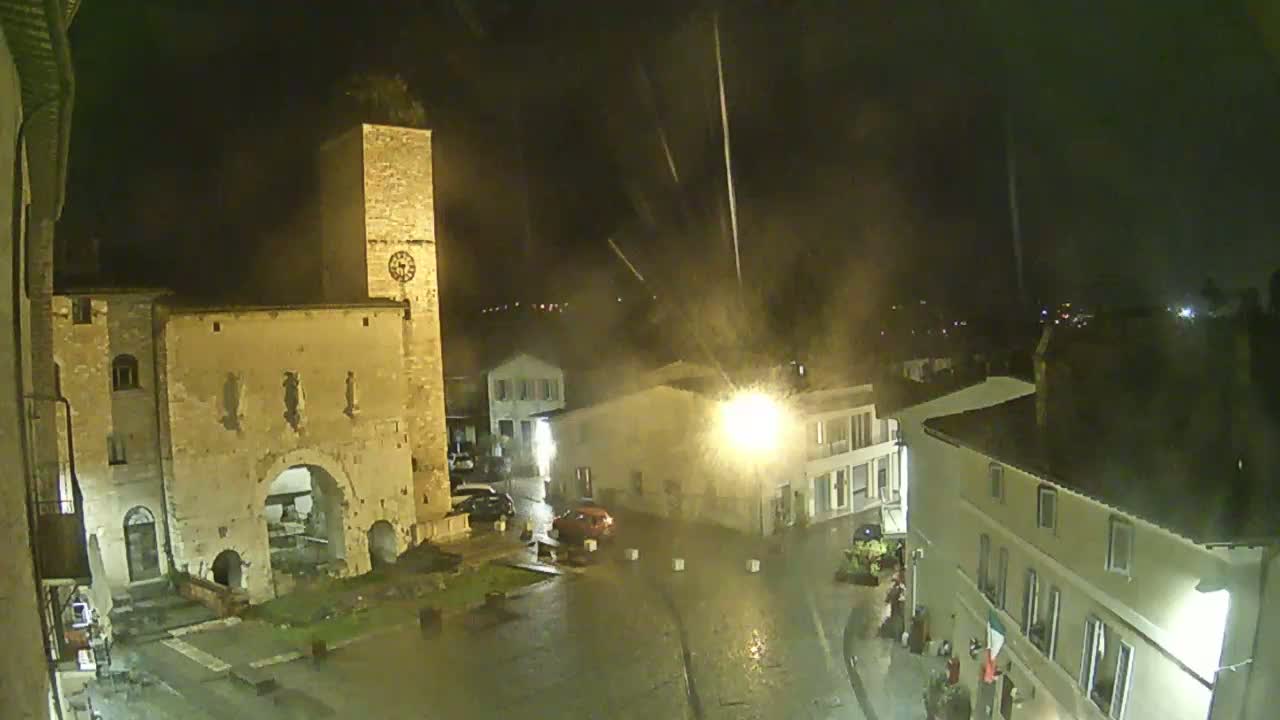 A rain-soaked town square at night is illuminated by streetlights, featuring an ancient stone gateway and clock tower under a dark, stormy sky.