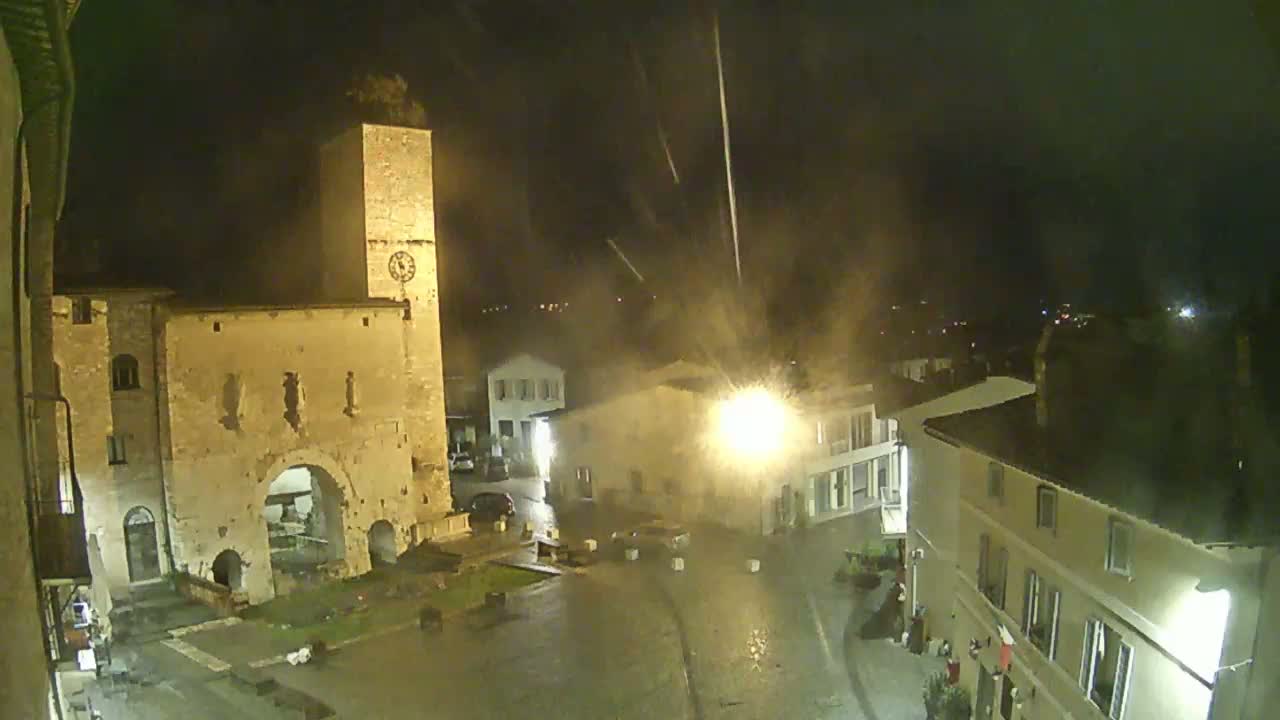 A nighttime view of a town square with old stone buildings, a tall clock tower, and rain falling.