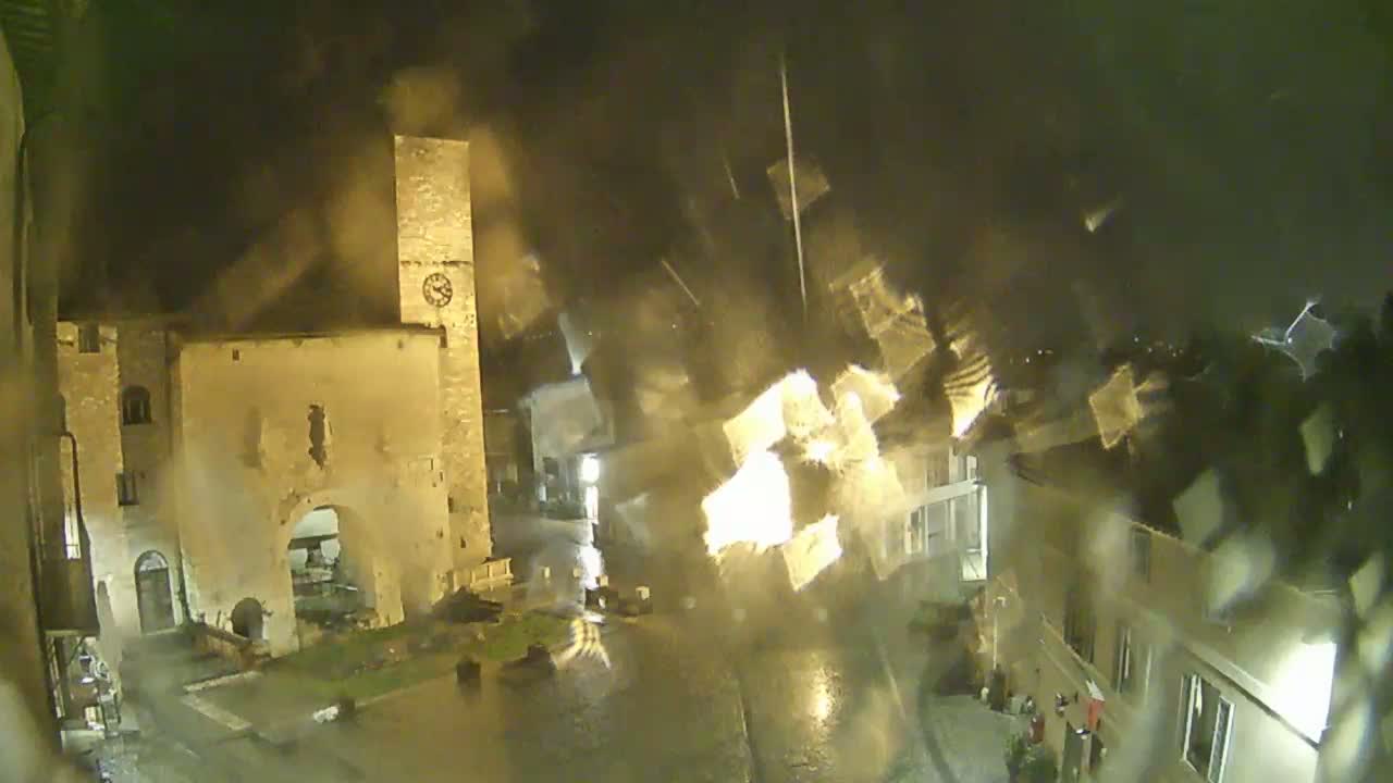 A nighttime view of a town square with a tall stone tower, viewed through rain-streaked glass during a heavy downpour.
