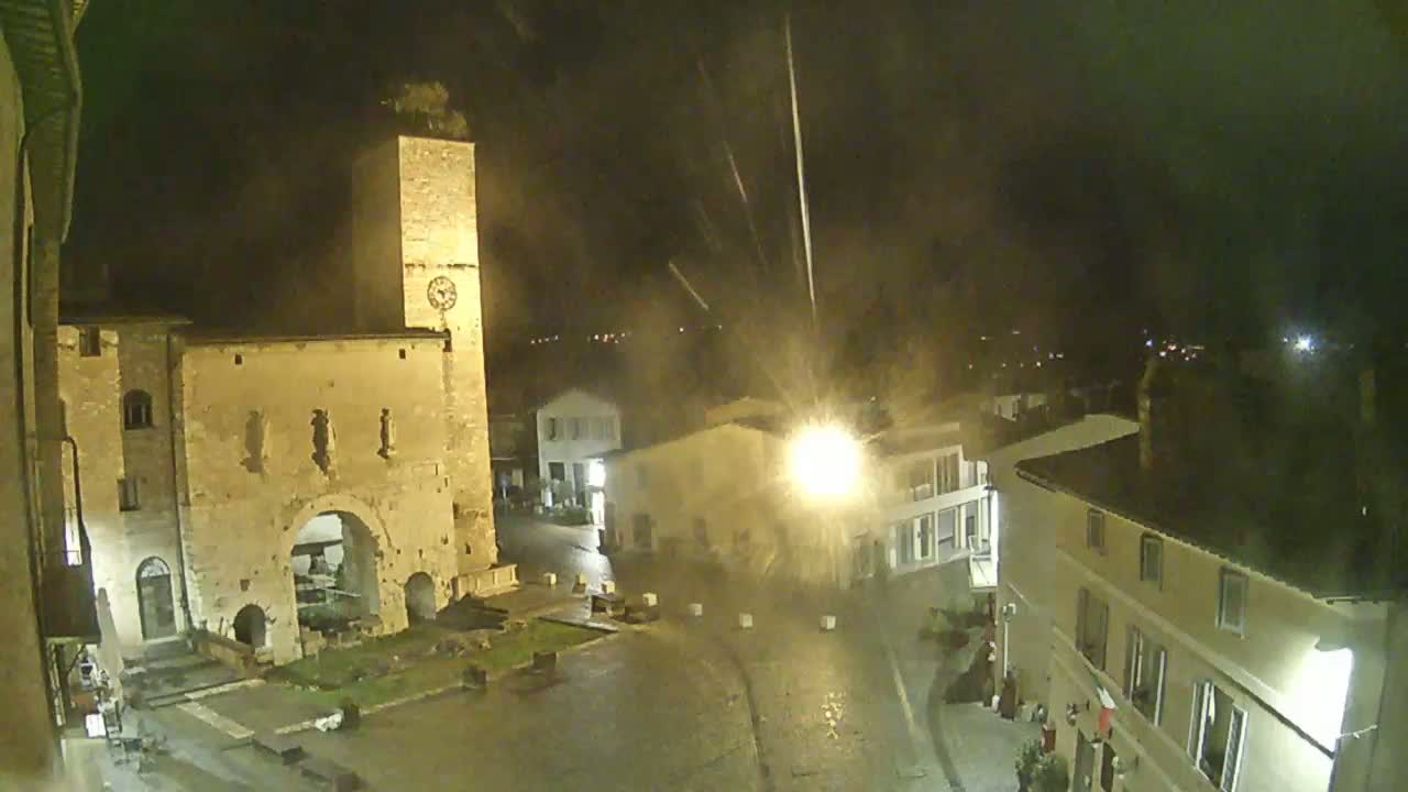A nighttime, rain-swept view of a town square dominated by a tall, illuminated stone tower and archway, with other buildings visible under streetlights.