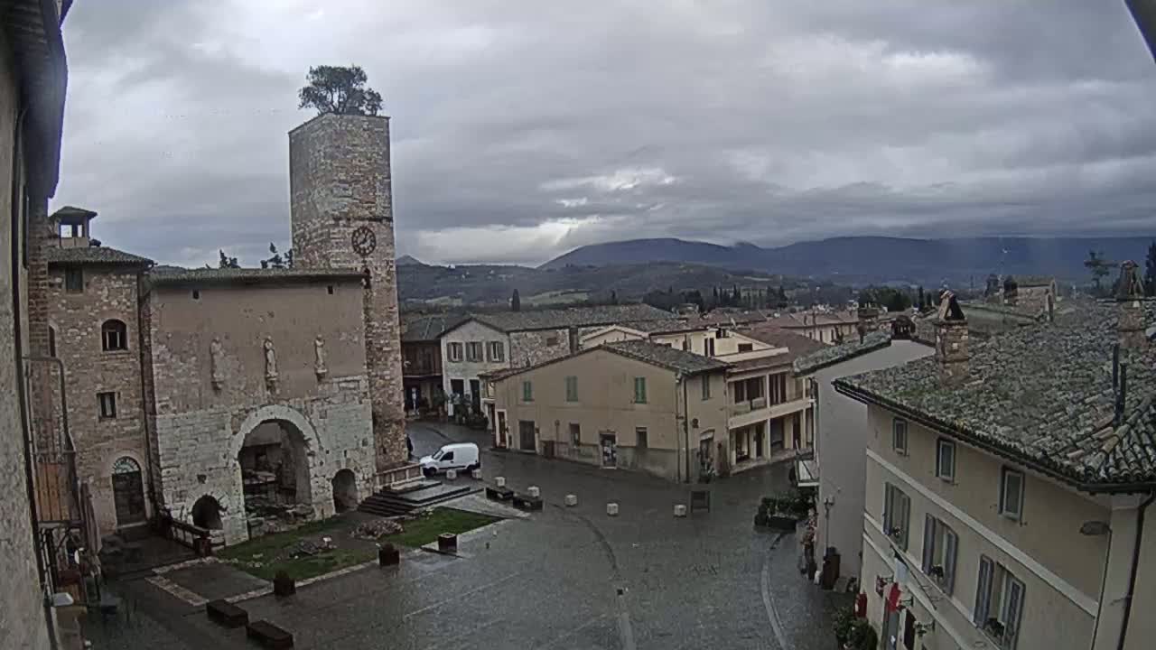 A town square in a small Italian town on a cloudy, overcast day, centered around an ancient stone building with a clock tower topped by a tree.