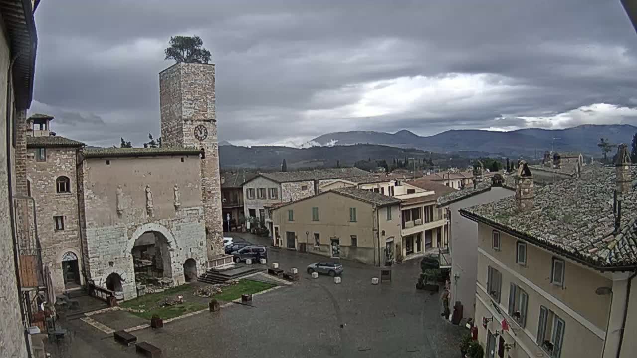 A town square in a small Italian town, under a cloudy sky, features an old stone building with a clock tower and a tree on top, surrounded by other buildings and a few cars.