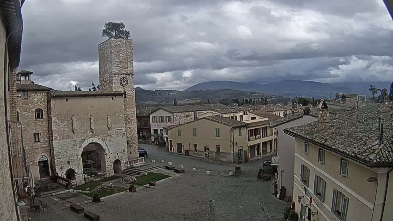 A town square in a historic Italian village under a cloudy sky, features a clock tower with a tree on top and surrounding buildings of stone.