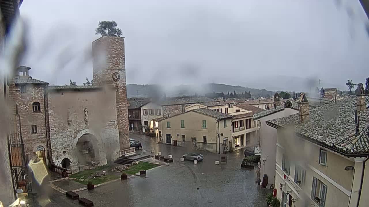 A rain-soaked town square in a hilltop town, featuring old stone buildings, a clock tower with a tree on top, and a few cars parked in the wet plaza.