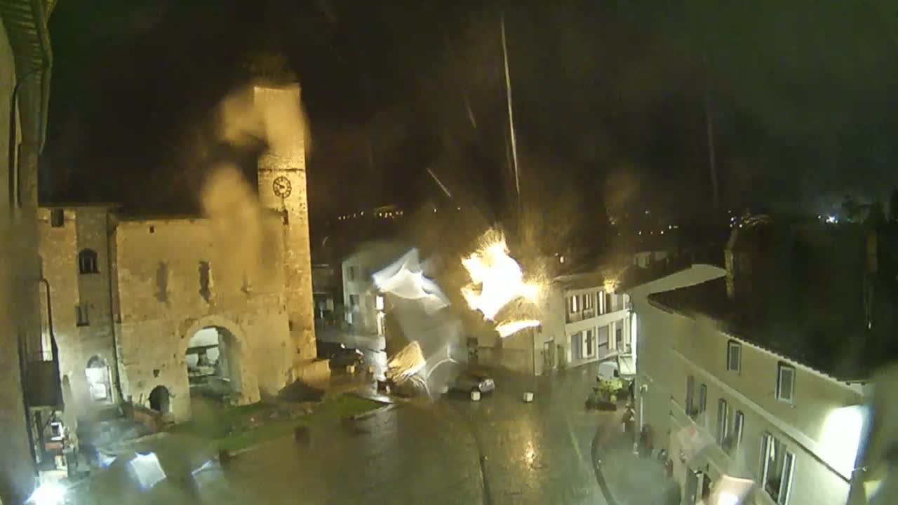 A nighttime view of a town square, illuminated by streetlights, showing an old stone archway and buildings under a heavy rain.