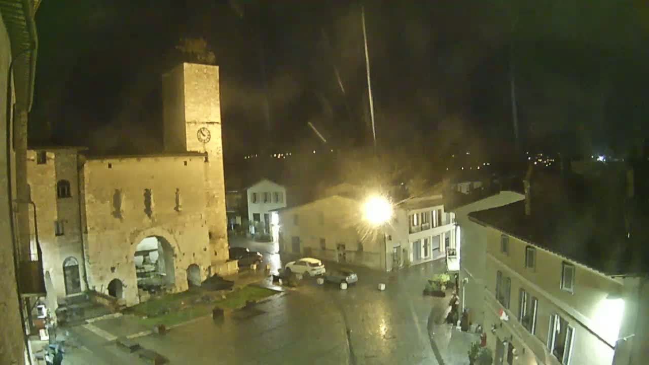 A rain-slicked town square at night is illuminated by streetlights, showing a medieval-looking stone building and other buildings under a dark, rainy sky.