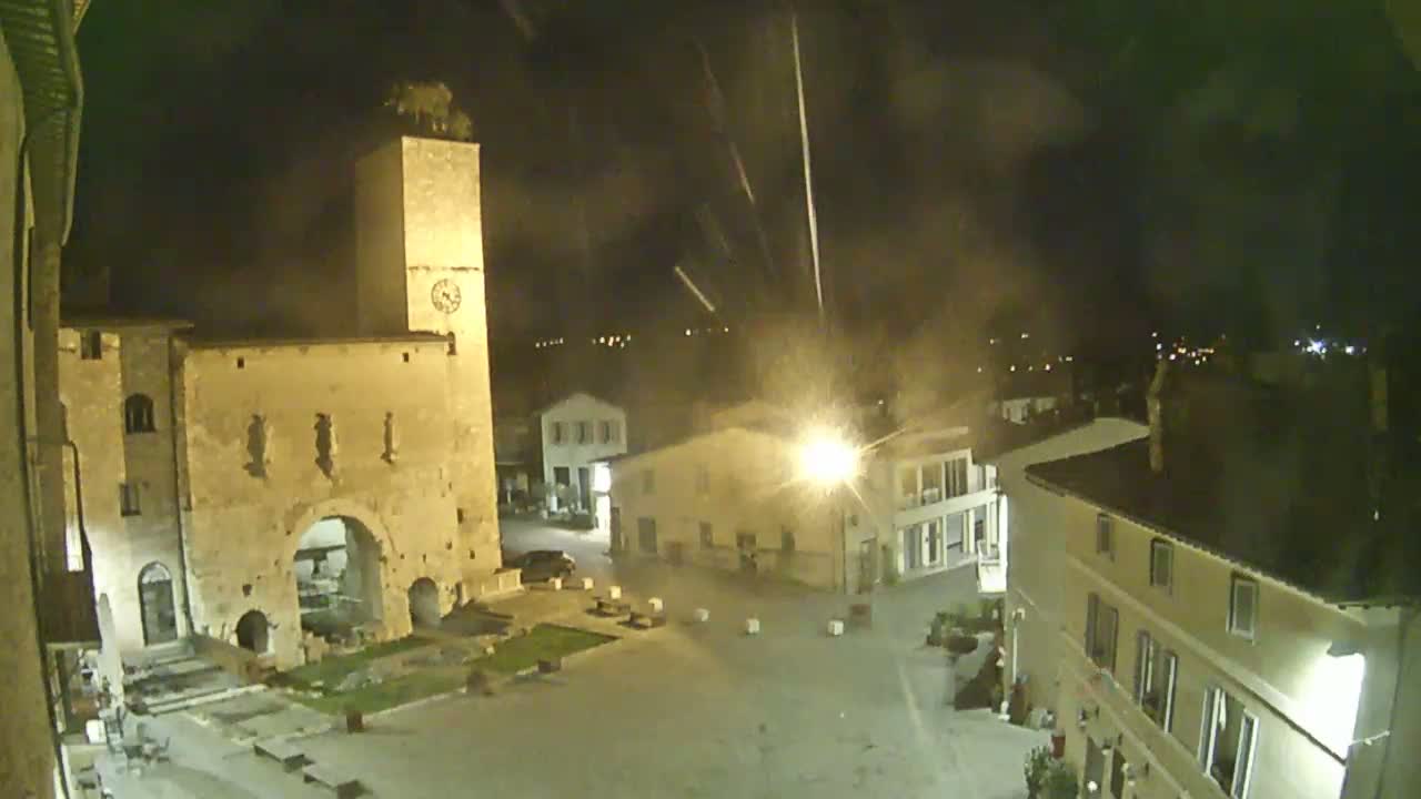 A nighttime, high-angle view of a town square with ancient-looking buildings, including a tall clock tower, under a slightly hazy sky.