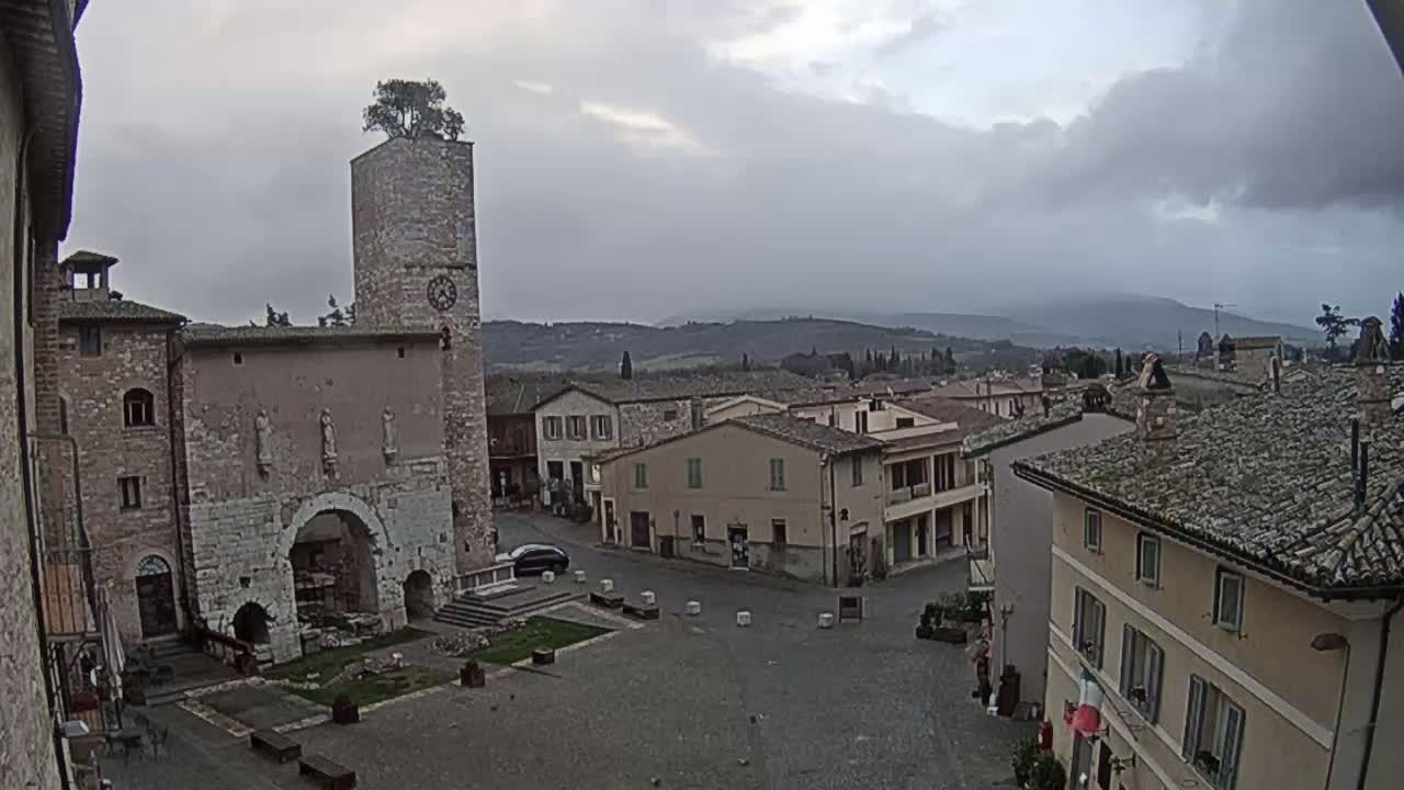 A town square in a small Italian town, under an overcast sky, is surrounded by stone buildings, one of which has a clock tower with a tree on top.