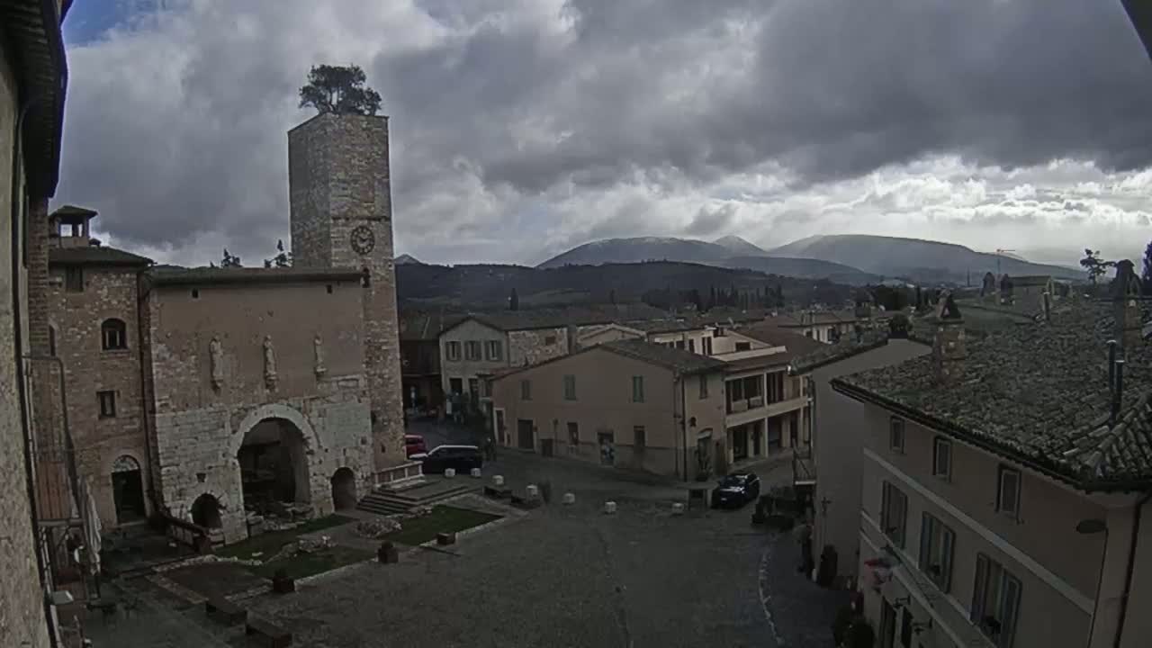A town square in a medieval-style village under a cloudy sky, features a tall stone tower with a tree on top, and mountains in the background.